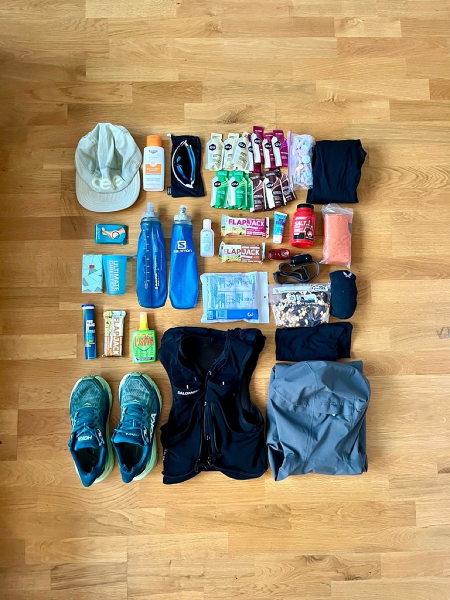 Flat lay of trail-running kit: shoes, hydration vest, flasks, gels and a cap on a wooden floor