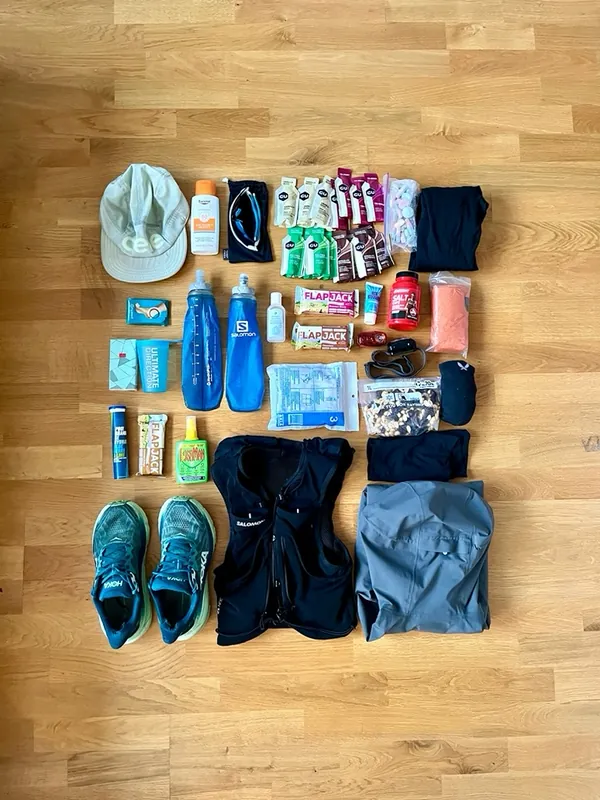 Flat lay of trail-running kit: shoes, hydration vest, flasks, gels and a cap on a wooden floor