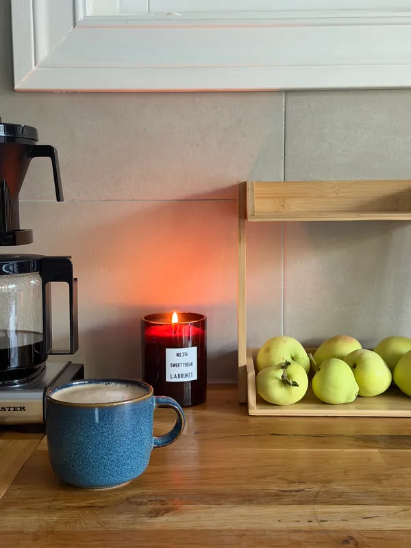 A lit L:A Bruket Sweet Tobak candle on a wooden kitchen counter, beside a blue coffee mug and green apples