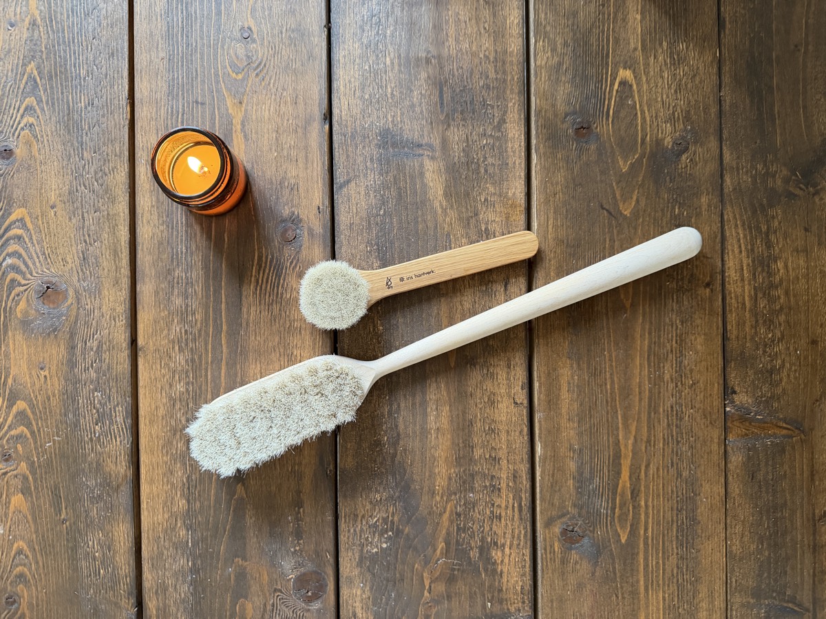 Two natural-bristle body brushes and a lit amber candle on dark wood
