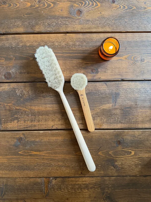 Two natural-bristle body brushes and a lit amber candle on dark wood