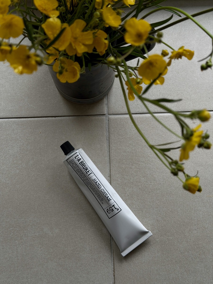 A hand cream tube on a tiled floor beside a vase of yellow buttercups