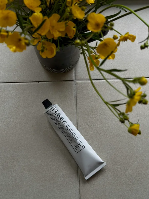 A hand cream tube on a tiled floor beside a vase of yellow buttercups