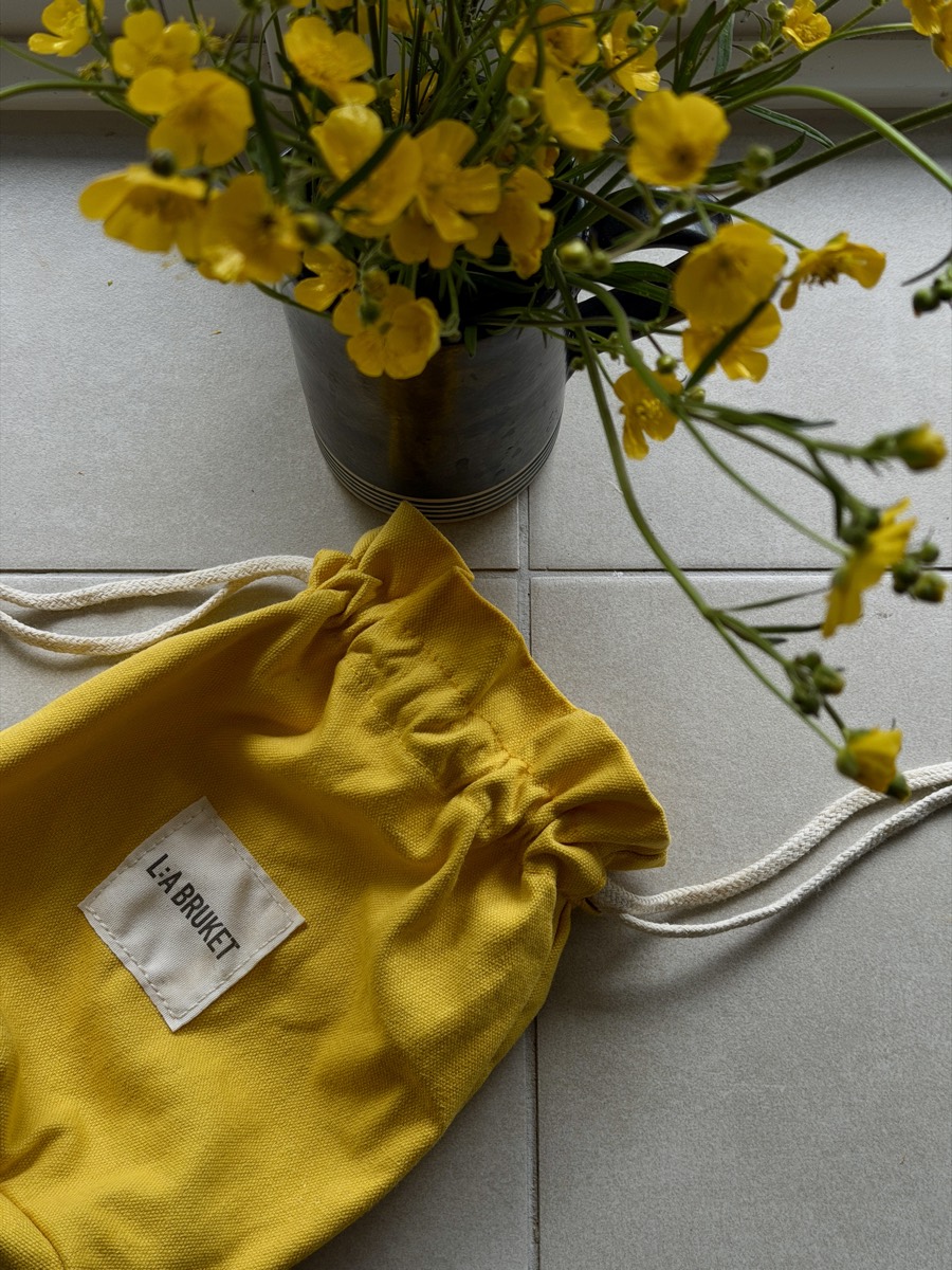A yellow cotton drawstring pouch on a tiled floor beside yellow buttercups