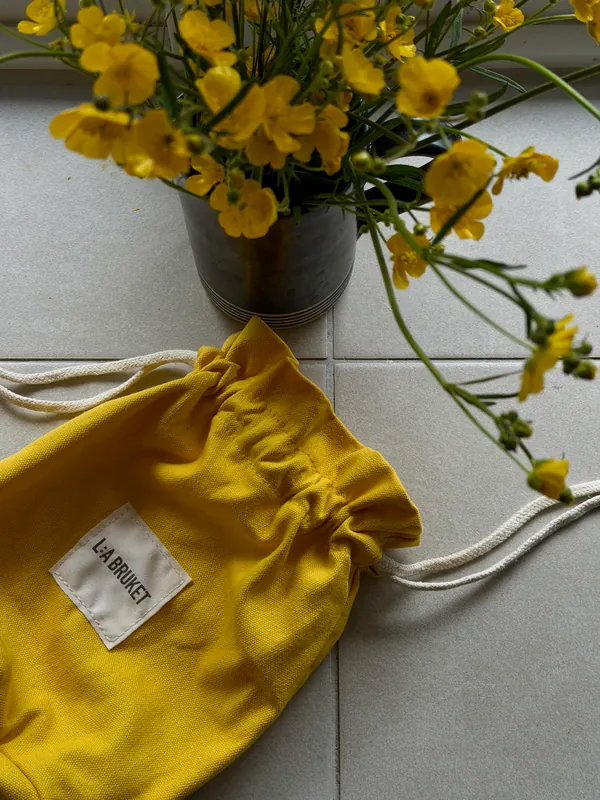 A yellow cotton drawstring pouch on a tiled floor beside yellow buttercups