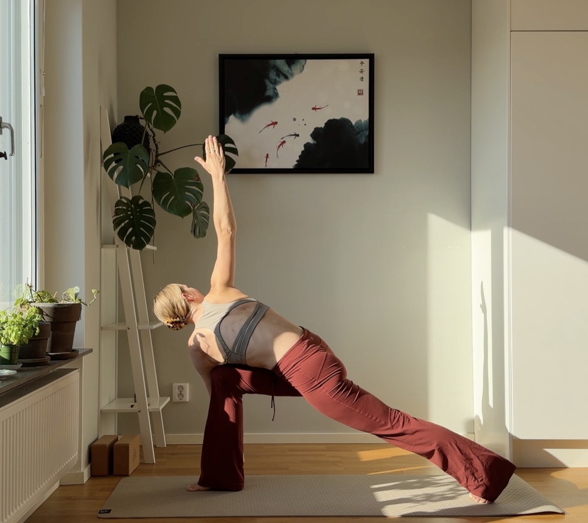 Camilla in a side-angle yoga pose in a sunlit Stockholm apartment, wearing red flared trousers