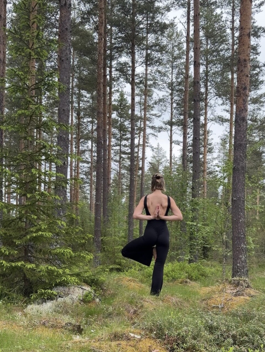 Camilla in tree pose seen from behind, among tall pines in a Swedish forest