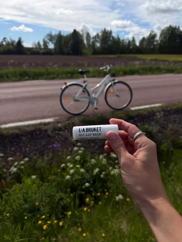 A hand holding an L:A Bruket lip balm by a country road, bicycle and summer meadow behind