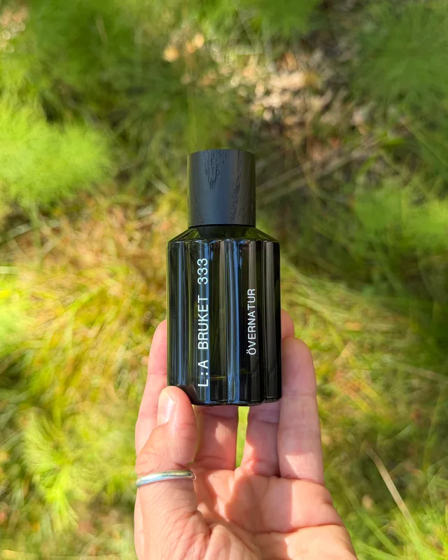 A hand holding an L:A Bruket Övernatur fragrance bottle close up, soft green forest floor behind
